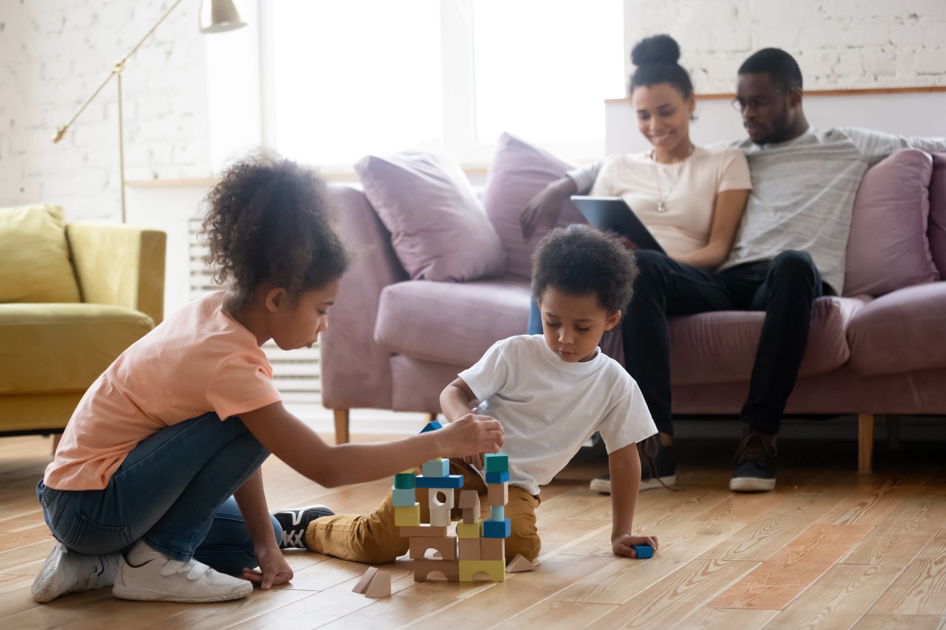 Children playing with blocks on the floor, parents on the couch in the background.