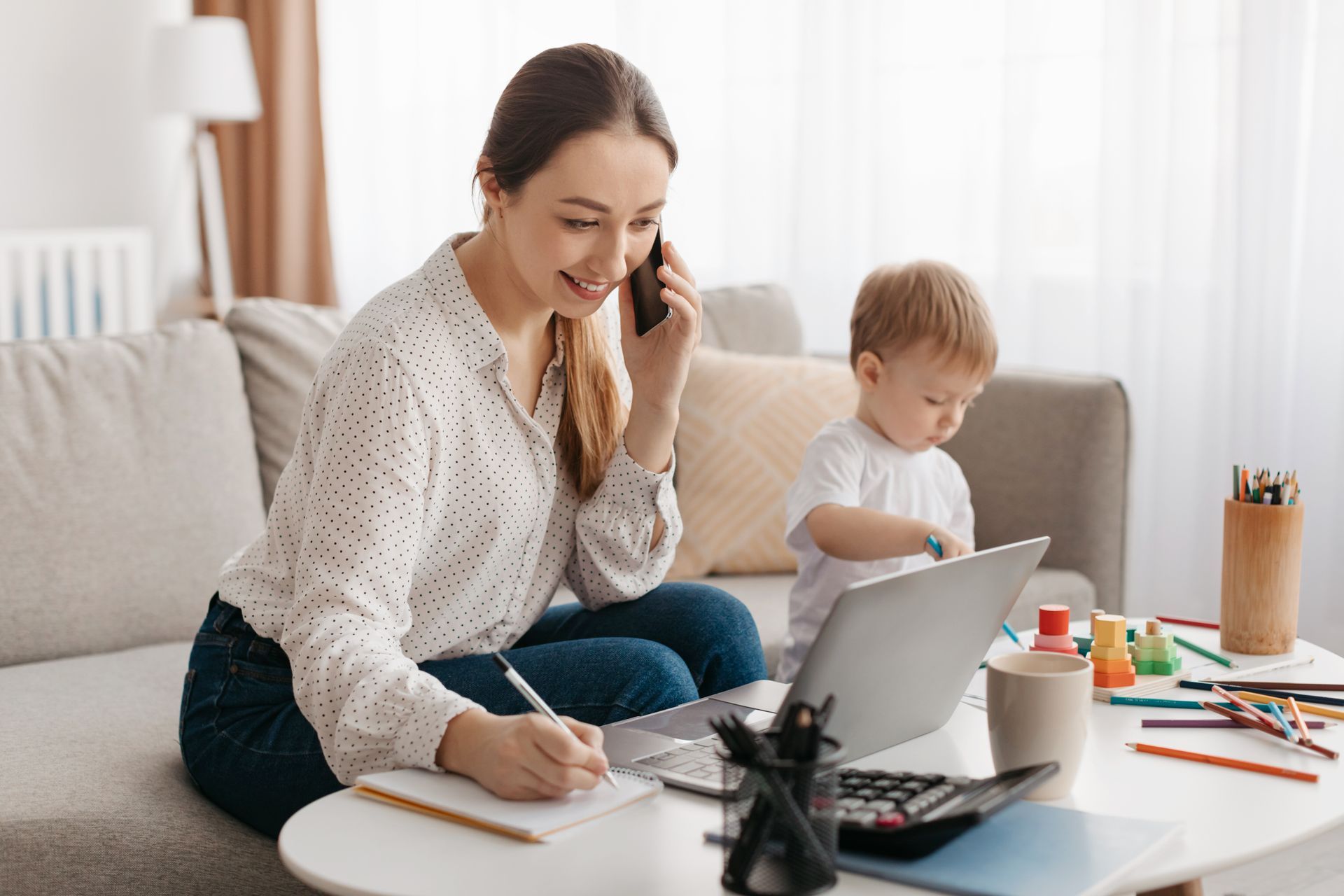 Woman working from home on the phone and laptop, taking notes, while a toddler plays nearby on the couch.