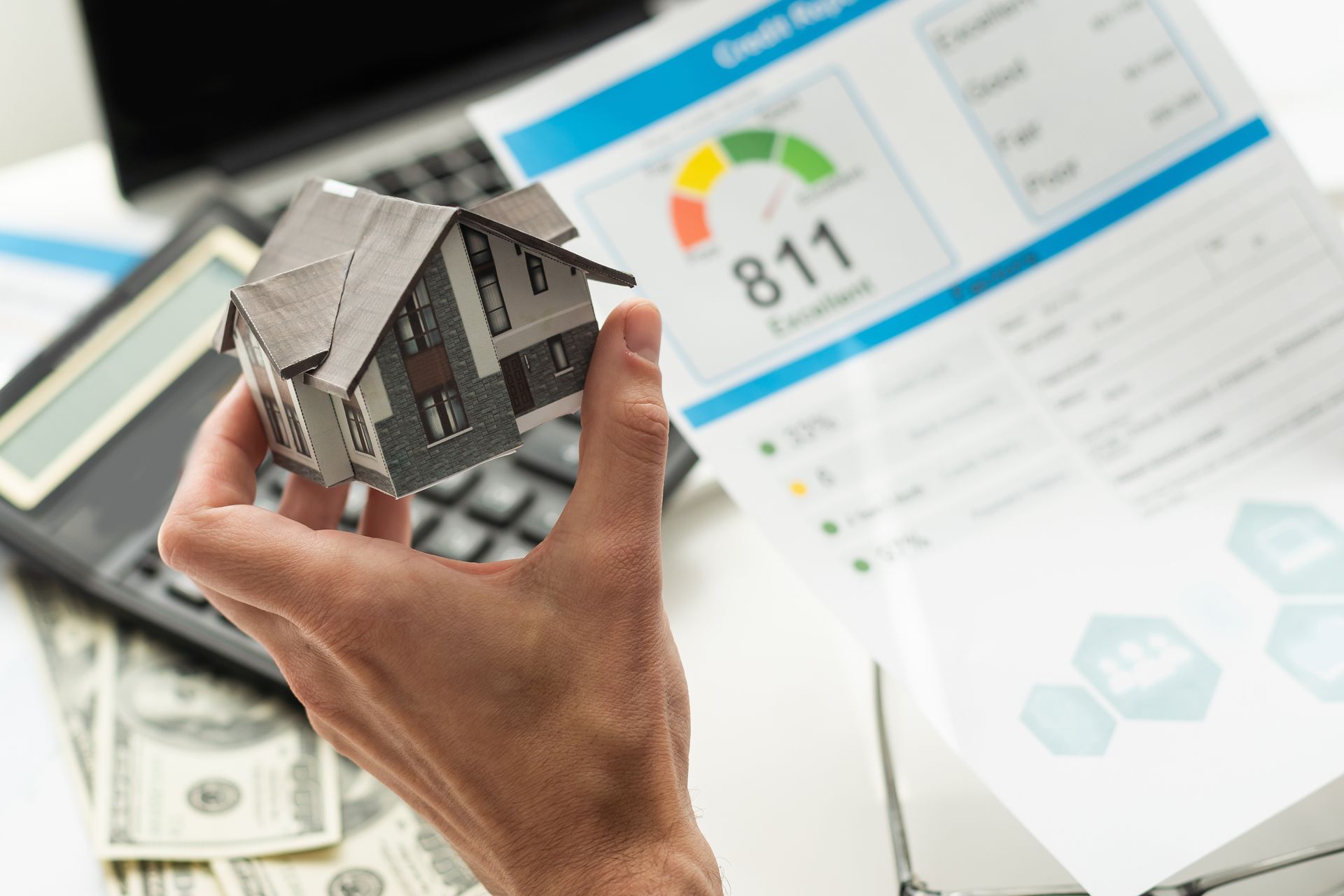 Hand holding a miniature house, with credit report and money visible in the background.