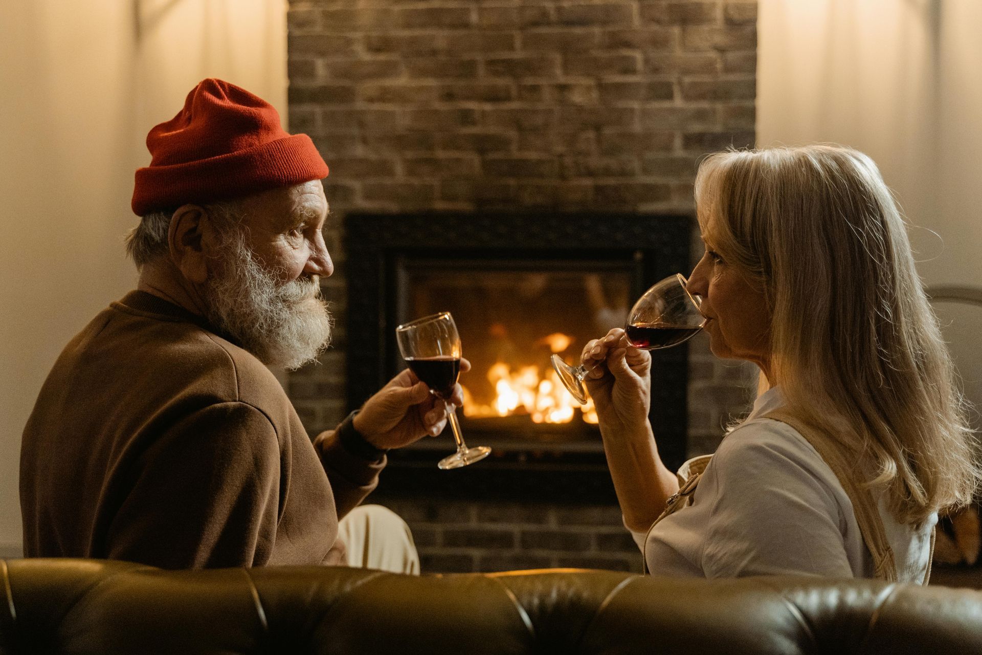 Couple toasting with wine by a fireplace; red hats, cozy setting.