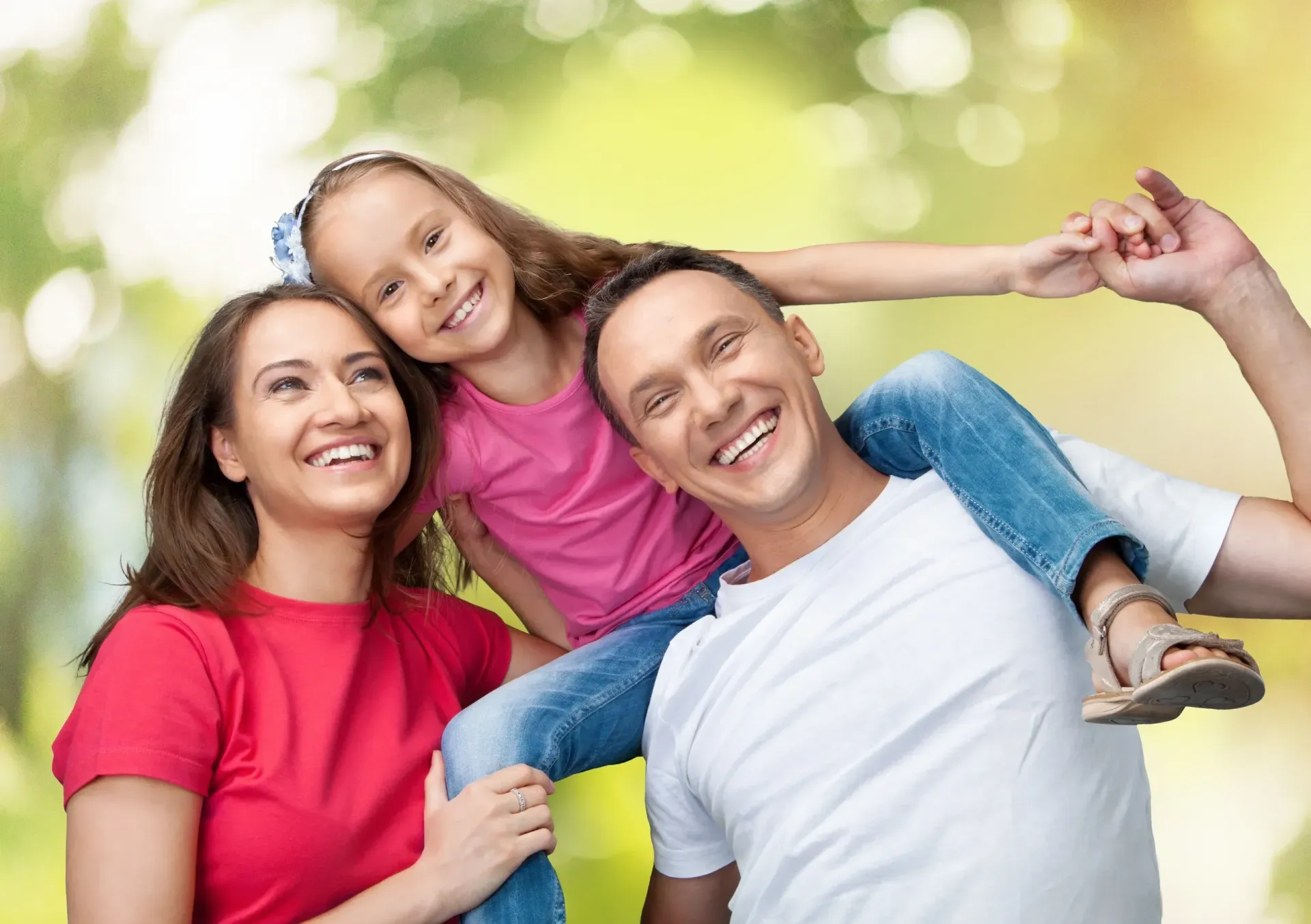 Family of three smiling outdoors; daughter rides on dad's shoulders, mom stands beside them. Green background.