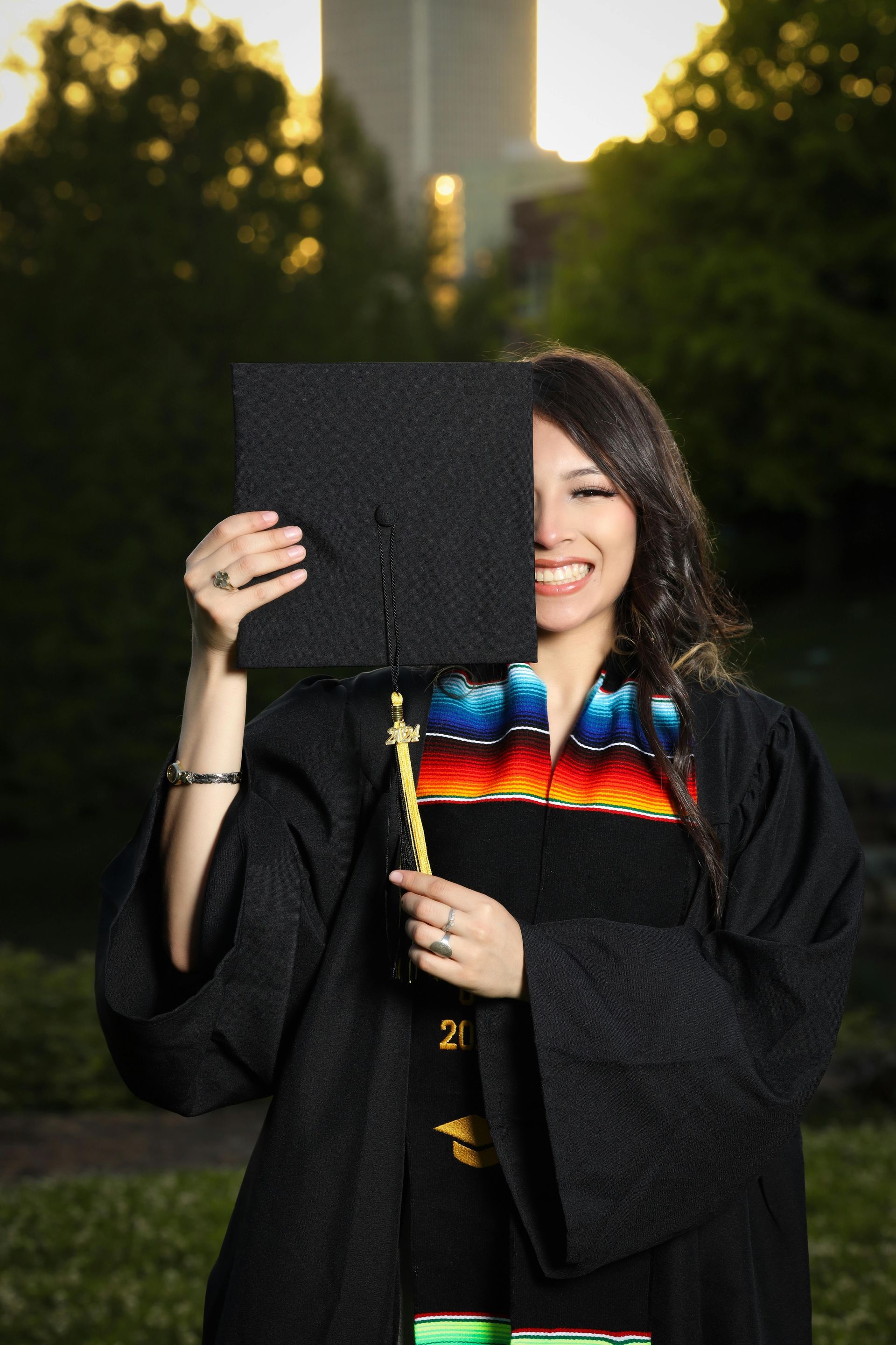 A smiling graduate in a black gown and decorated stole holds her graduation cap over one eye, standing outdoors.