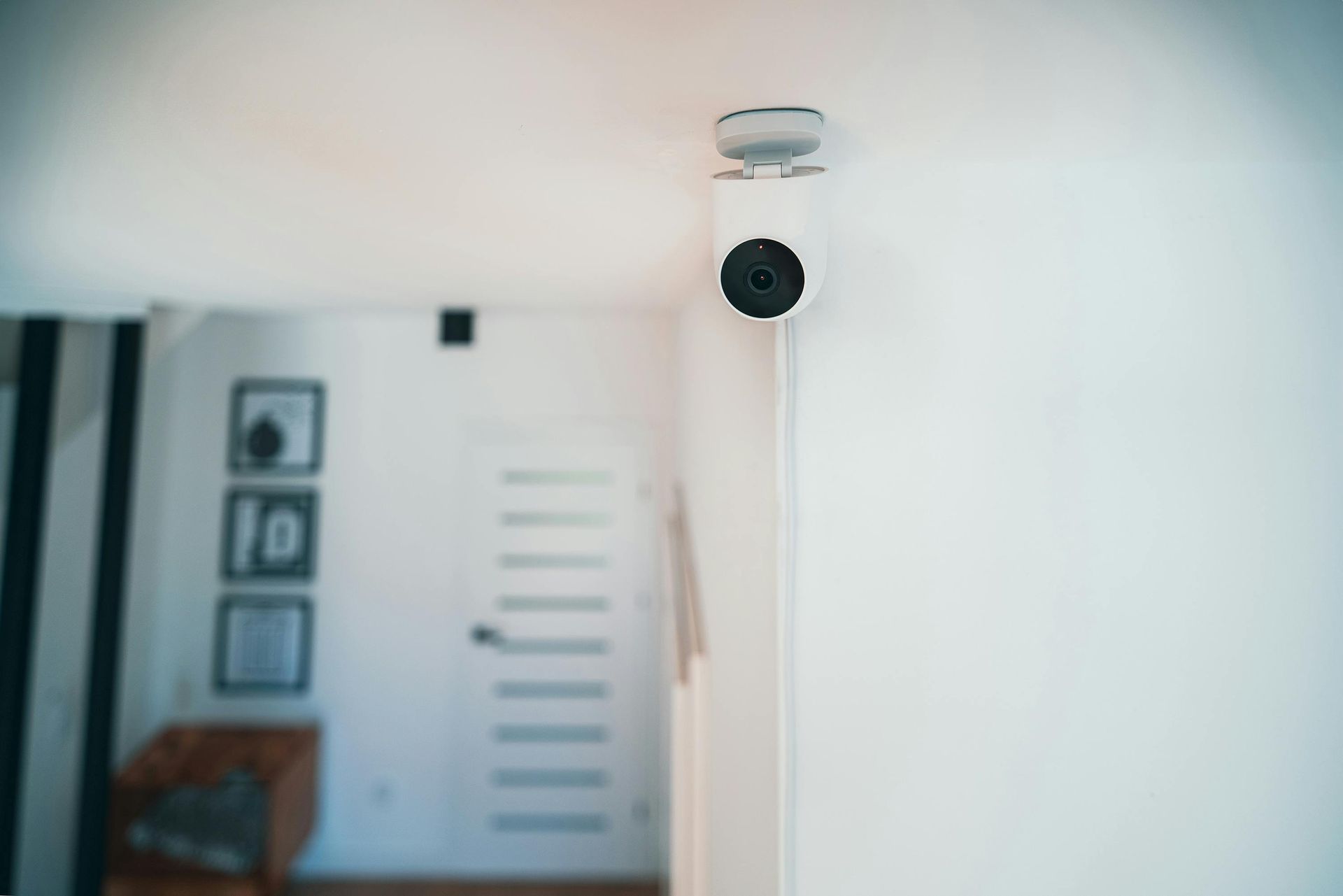 Security camera mounted on a white ceiling, pointed toward a hallway. The room has white walls and framed art.