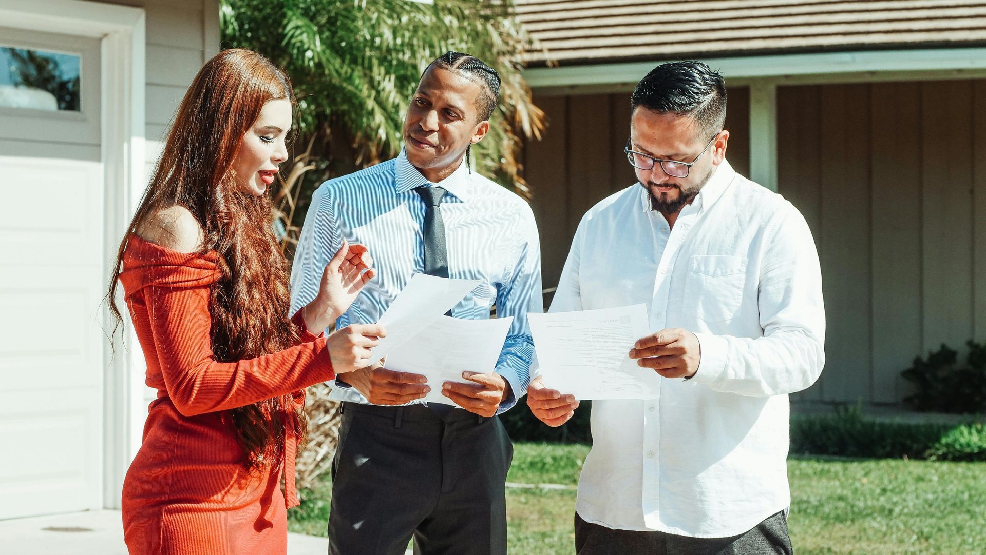 Three people reviewing documents outside a house. A woman in red dress gestures while two men in business attire examine papers.