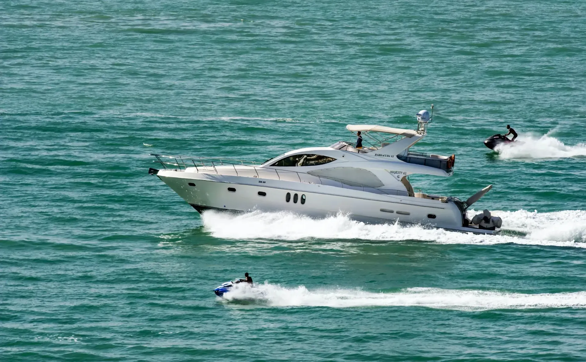 A white yacht cruises on turquoise water with two jet skis nearby, creating splashes.