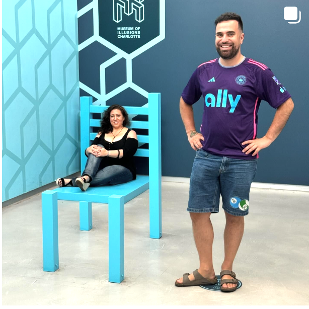 A man in a purple ally shirt stands next to a woman in a blue chair
