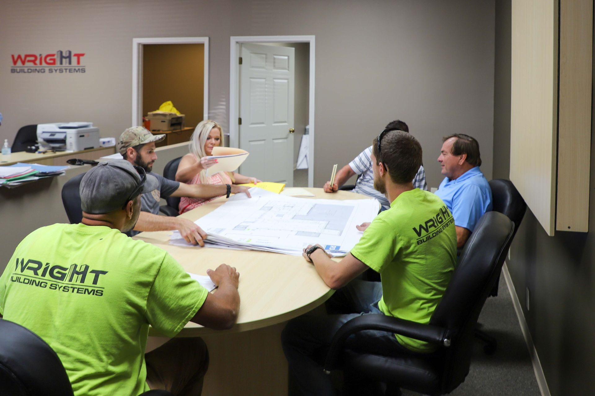 Six people around an oval table in a conference room, reviewing blueprints under a Wright Building Systems logo.