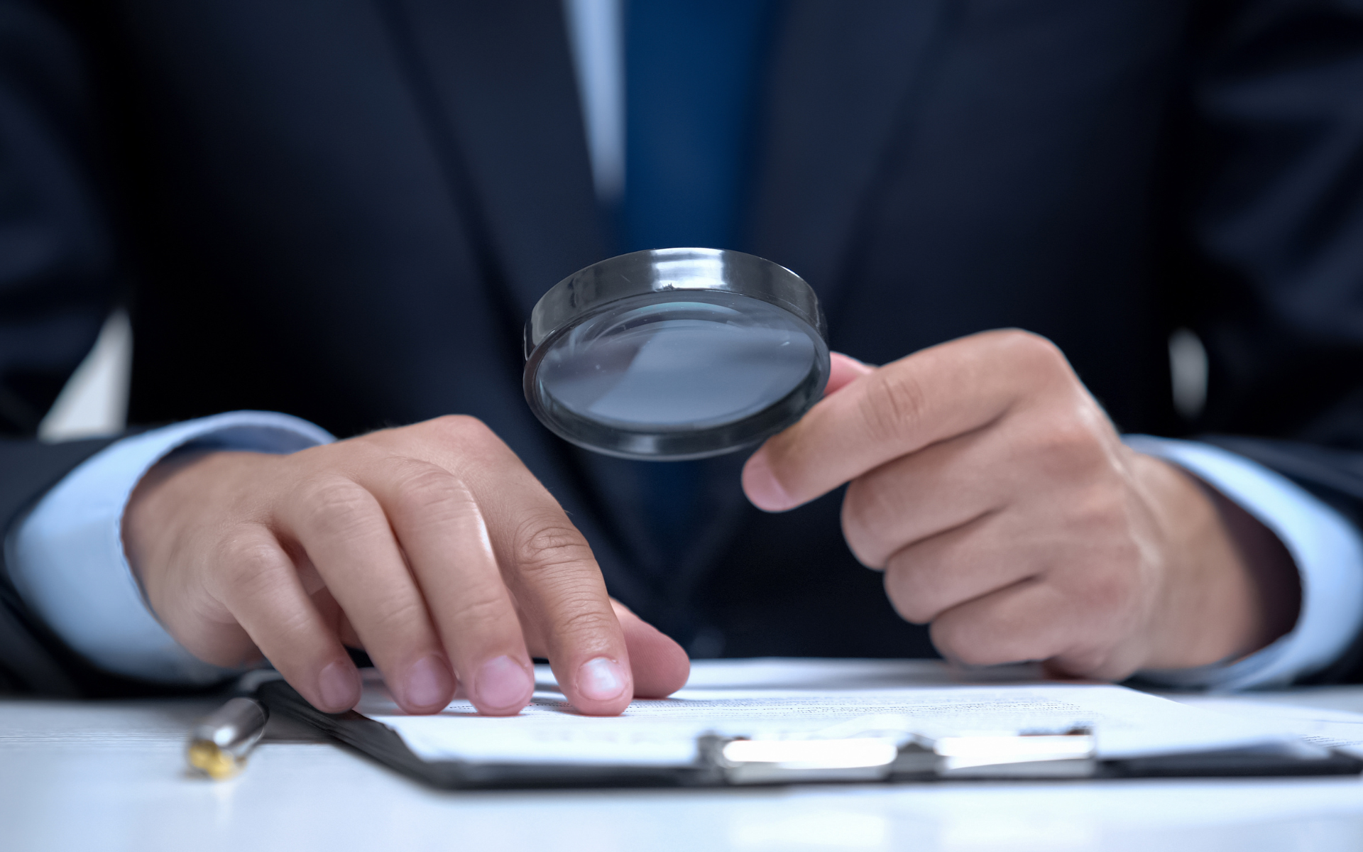 Person in suit examining document with magnifying glass.