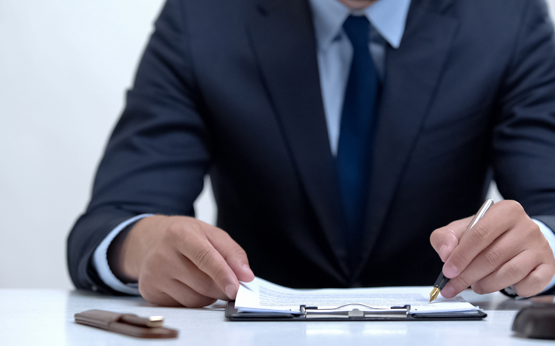 Man in suit reviewing paperwork at a desk, holding a pen.