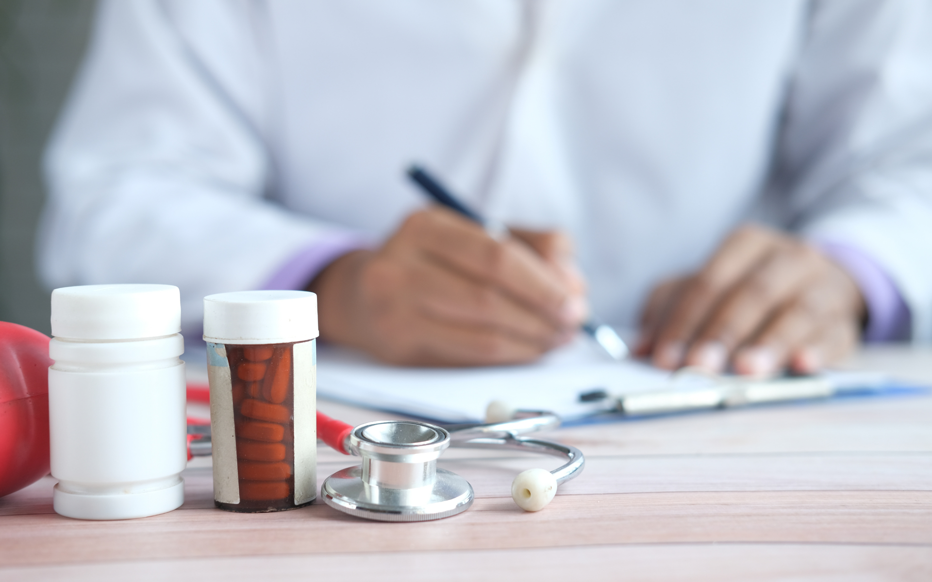Doctor writing on clipboard; stethoscope, pill bottles, and a heart are on the table.