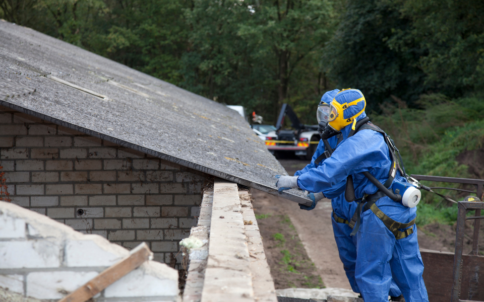 Two workers in blue hazmat suits remove roofing from a building, safety gear visible.