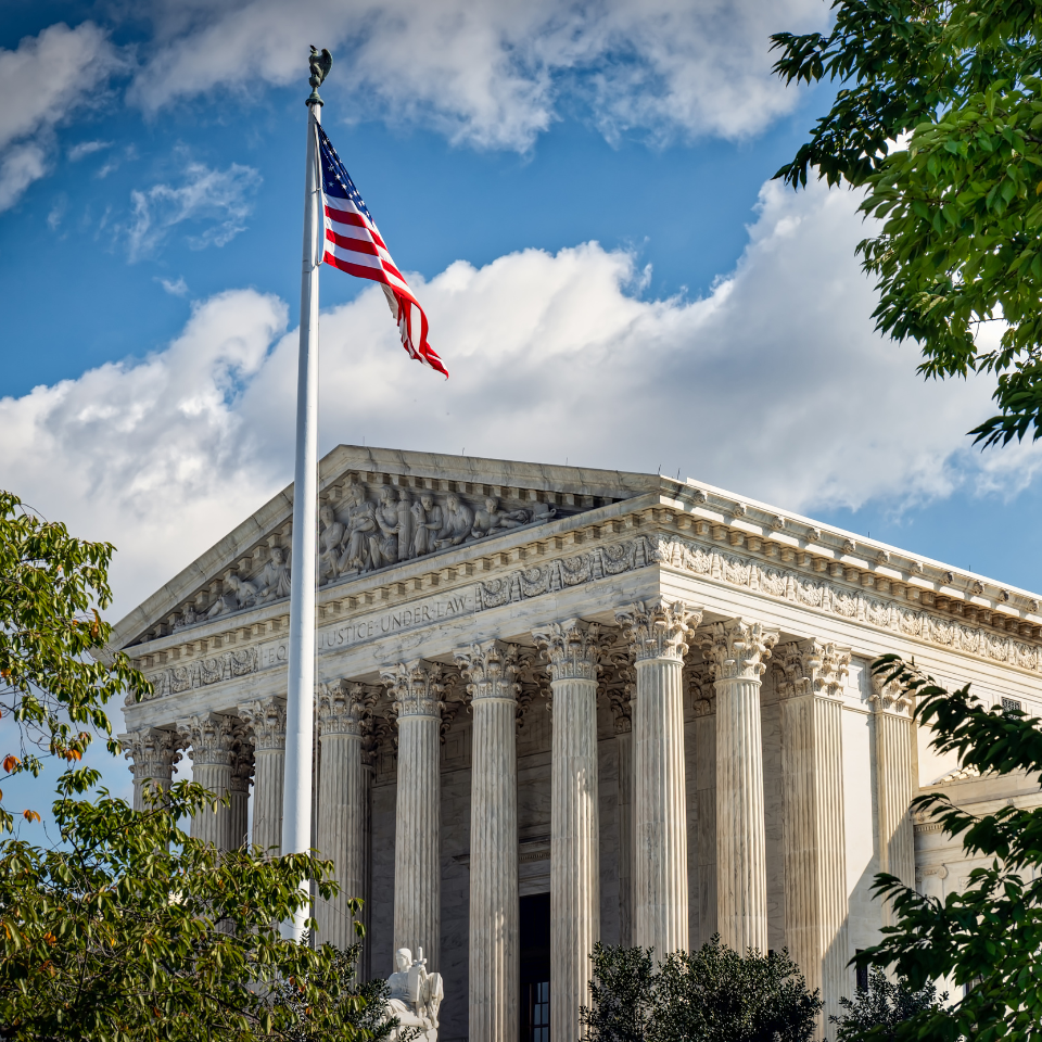 U.S. Supreme Court building with American flag flying in front of a blue sky and trees.