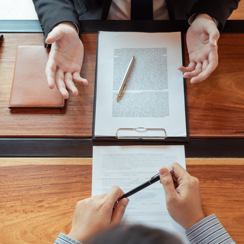 Two people at a wooden table reviewing documents, one gesturing with hands, the other holding a pen.