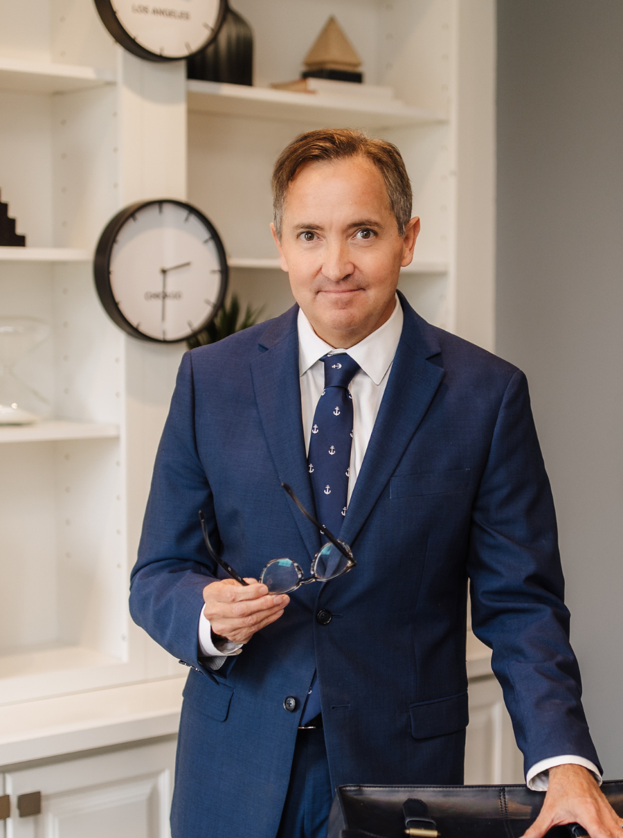 Man in a blue suit holding glasses, standing in an office with a bookshelf and a clock.