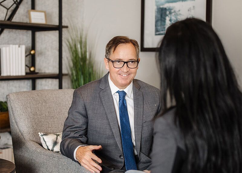 Man in a gray suit smiles, speaking to a woman. They sit in a room with a bookshelf.