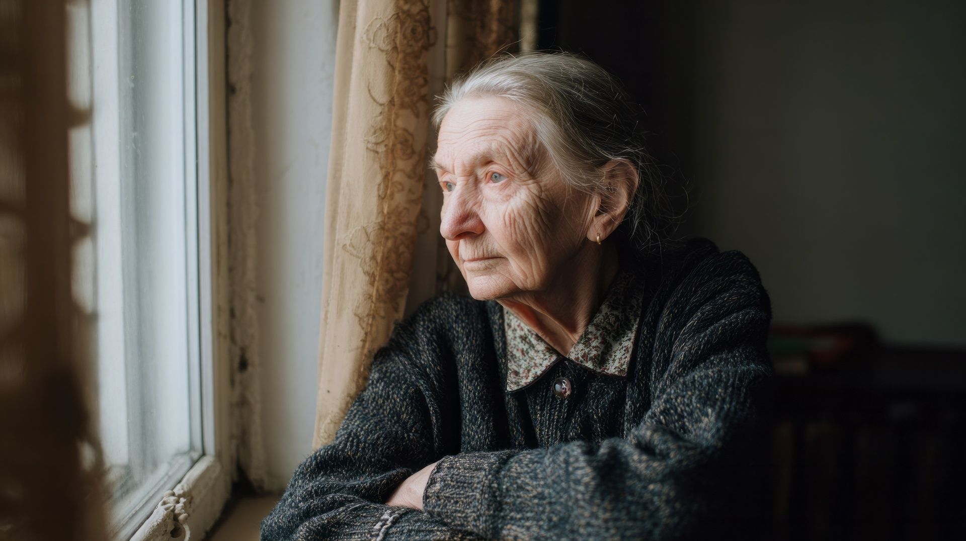 Elderly person gazing out a window, arms crossed, wearing a dark sweater in a dim room.