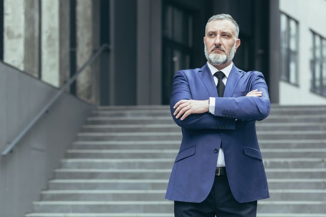 Man in blue suit, arms crossed, stands in front of building entrance.