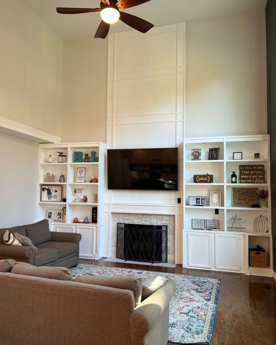 Living room with fireplace, built-in shelves, TV, and couches. White walls, dark wood floors, and a ceiling fan.