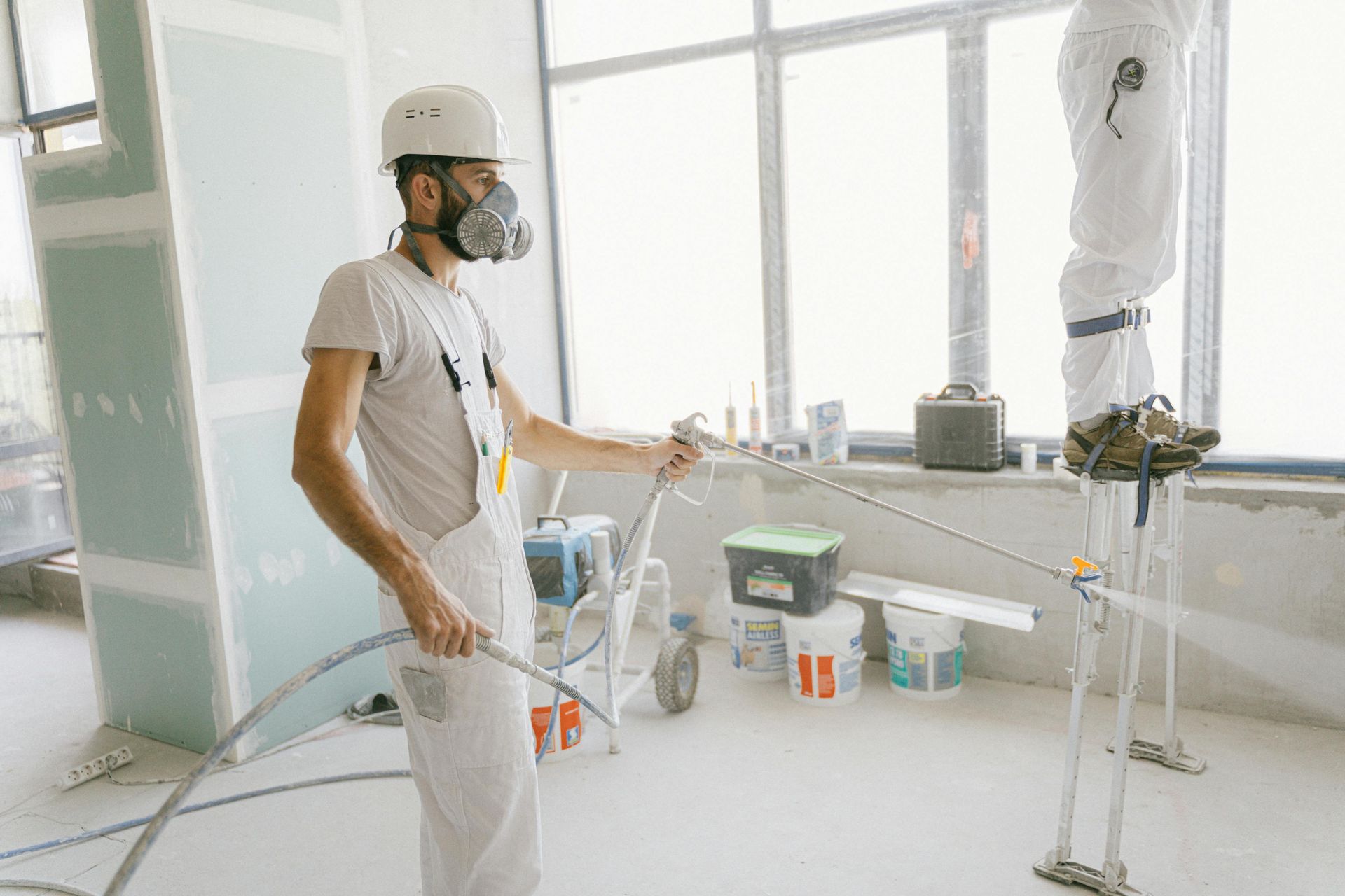 Man in respirator spraying paint in a construction site with another figure on stilts near a window.