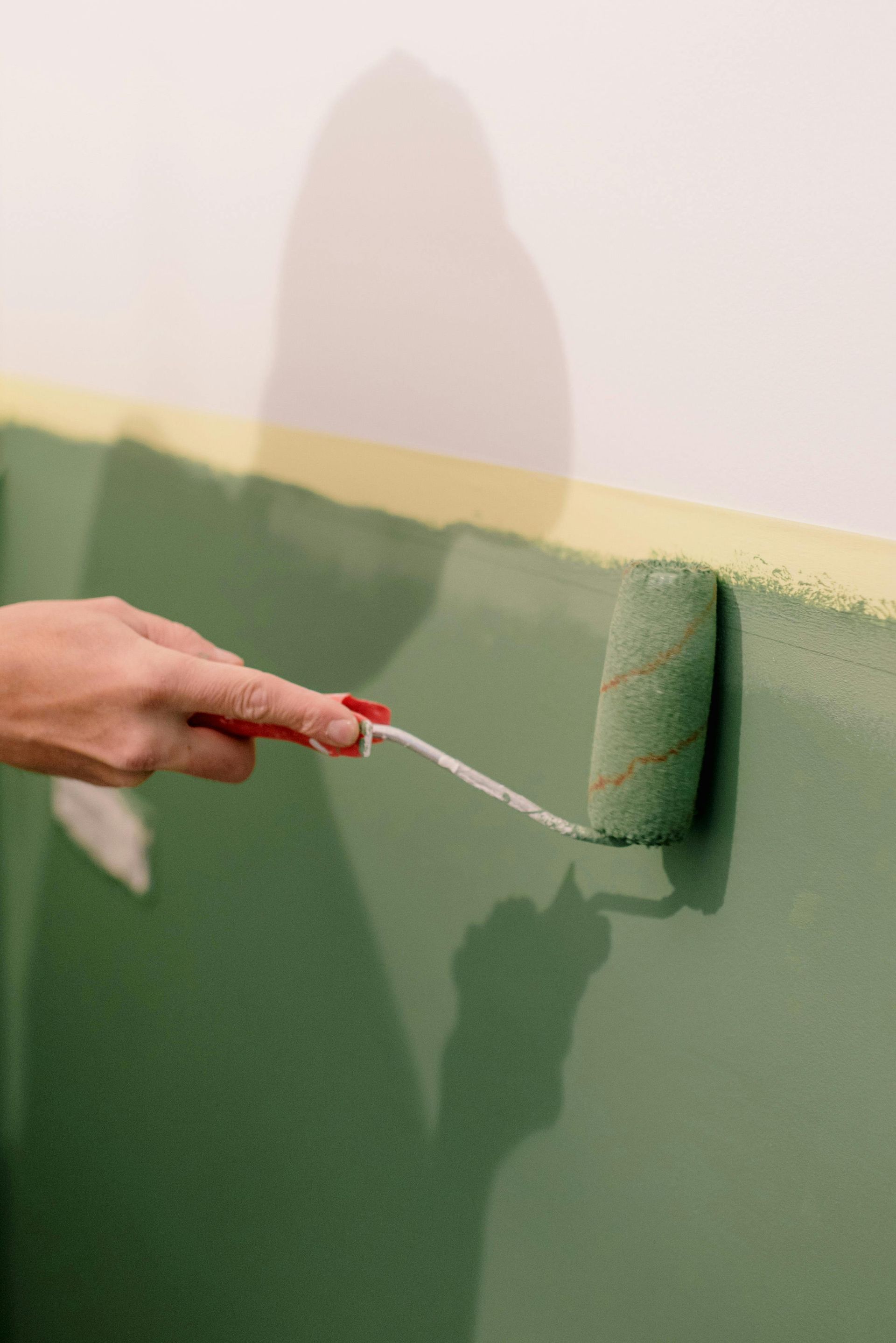 Hand using a paint roller to paint a wall green, with a white upper section and paint edge visible