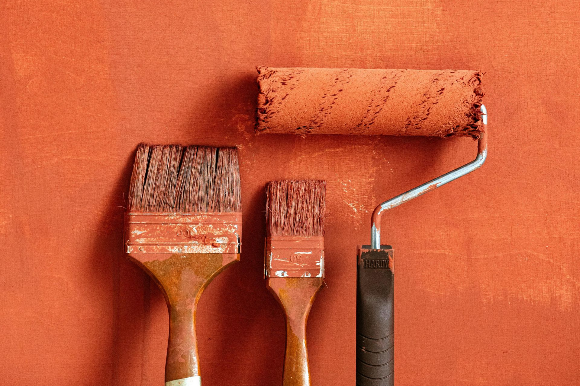 Three paint brushes and a roller with orange paint on a matching wall background