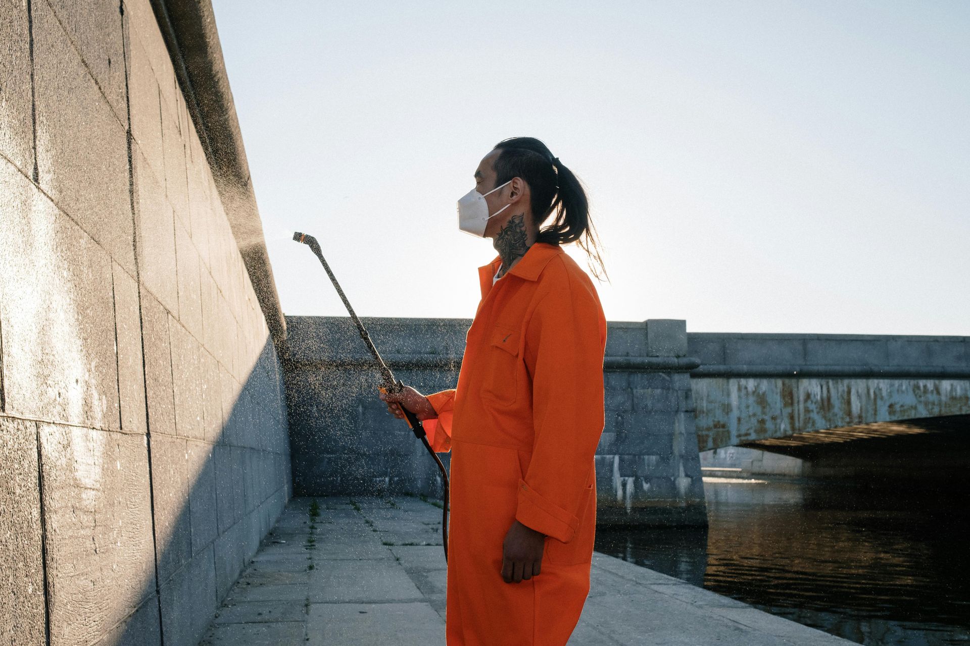 Person in orange jumpsuit holding a long pole beside a concrete wall near water, wearing a face mask.