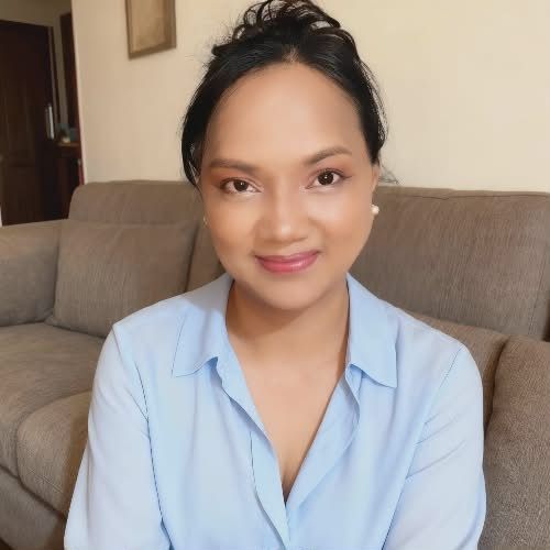 Smiling person seated on a beige couch, wearing a light blue blouse and white earrings.