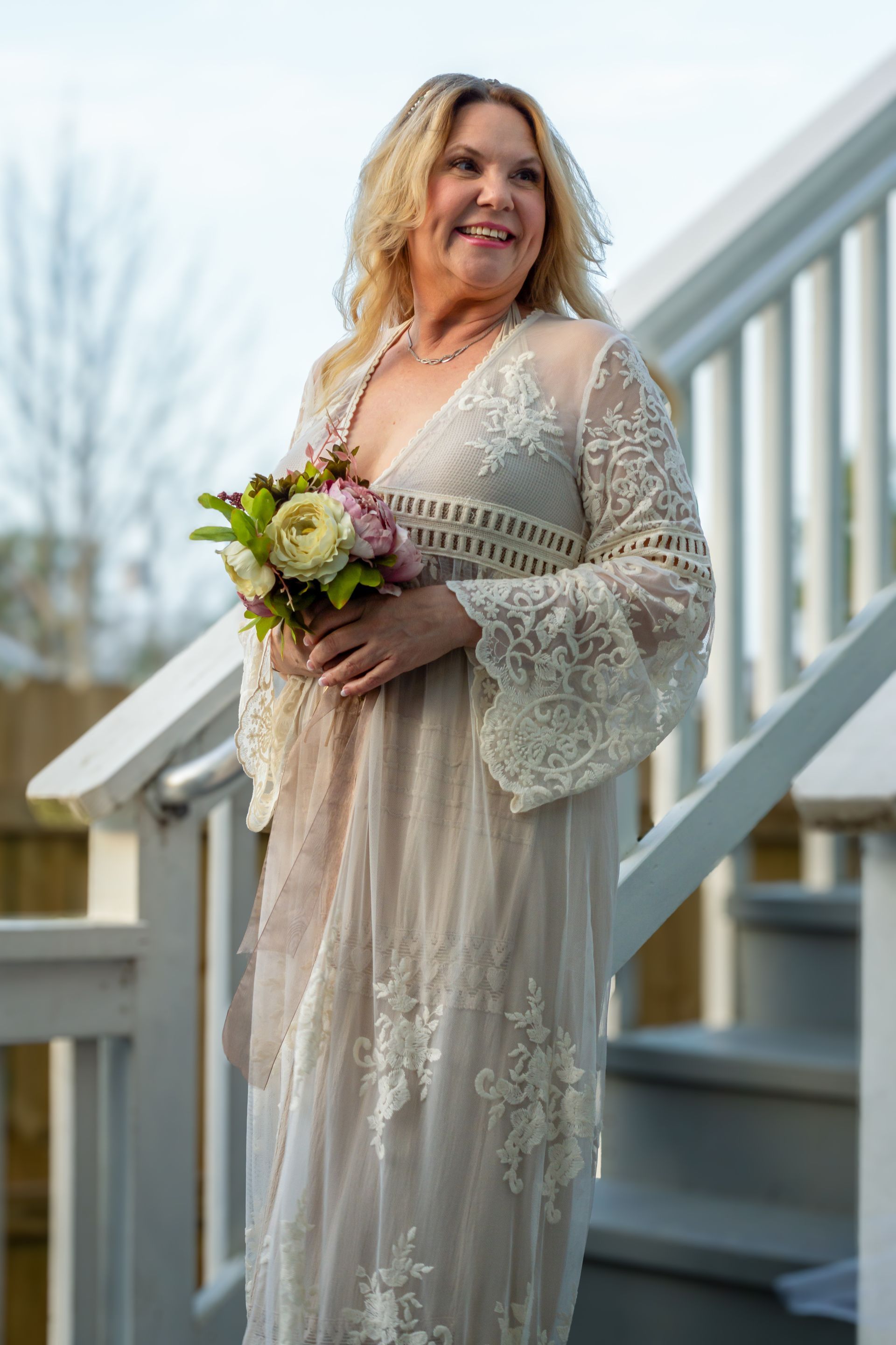 Woman in a lace dress holding a bouquet on outdoor stairs, smiling in soft daylight.
