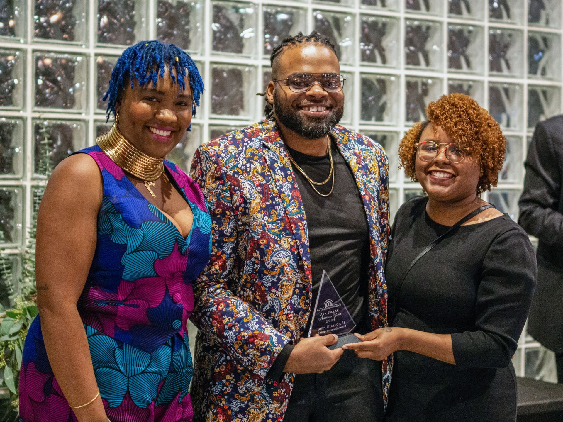 Three people smiling and holding a plaque in front of a glass block wall.