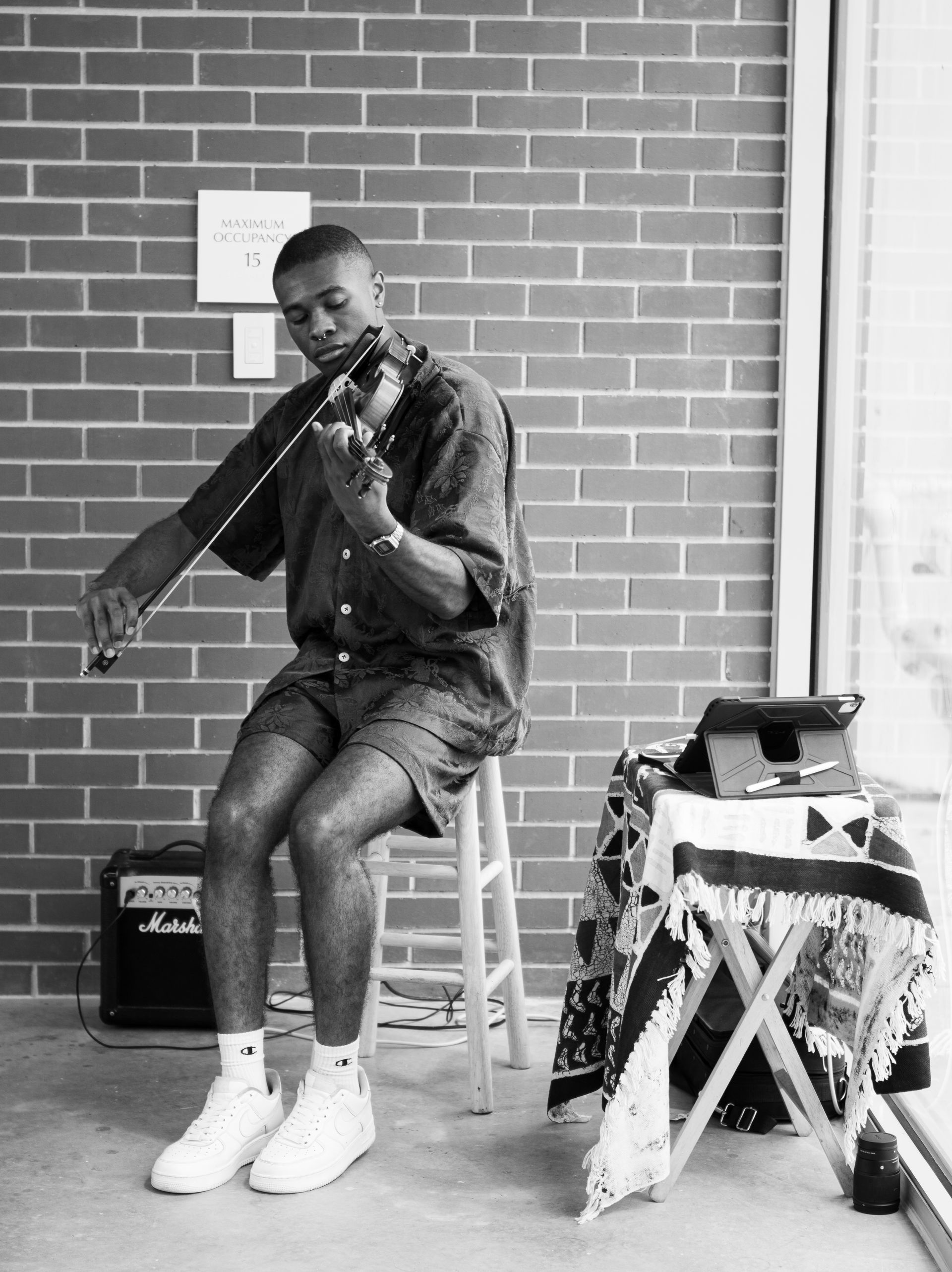 Person playing violin while seated on a stool beside a table in a brick-walled room