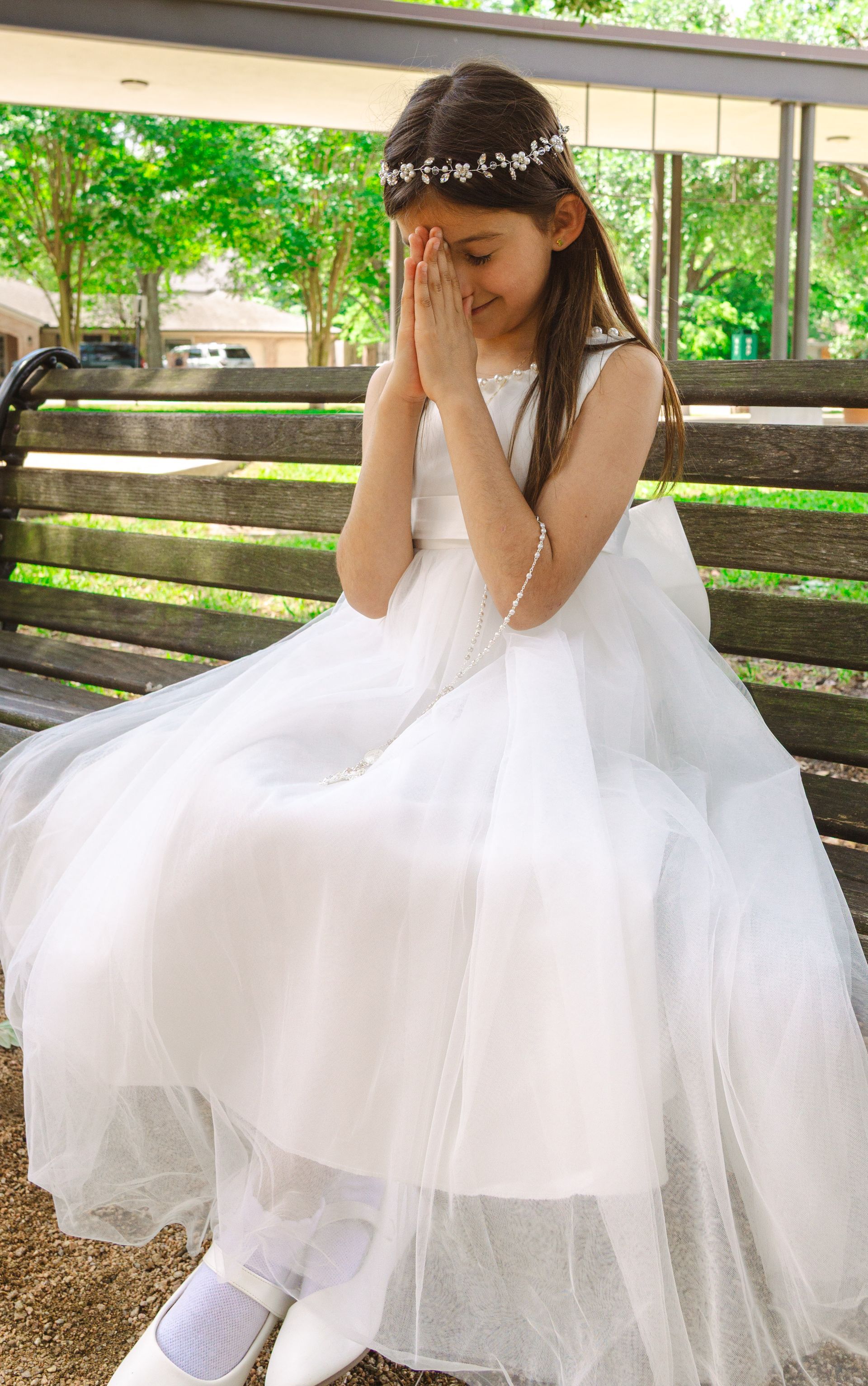 Young girl in a white dress and floral headband, sitting with hands together in a sunlit outdoor setting