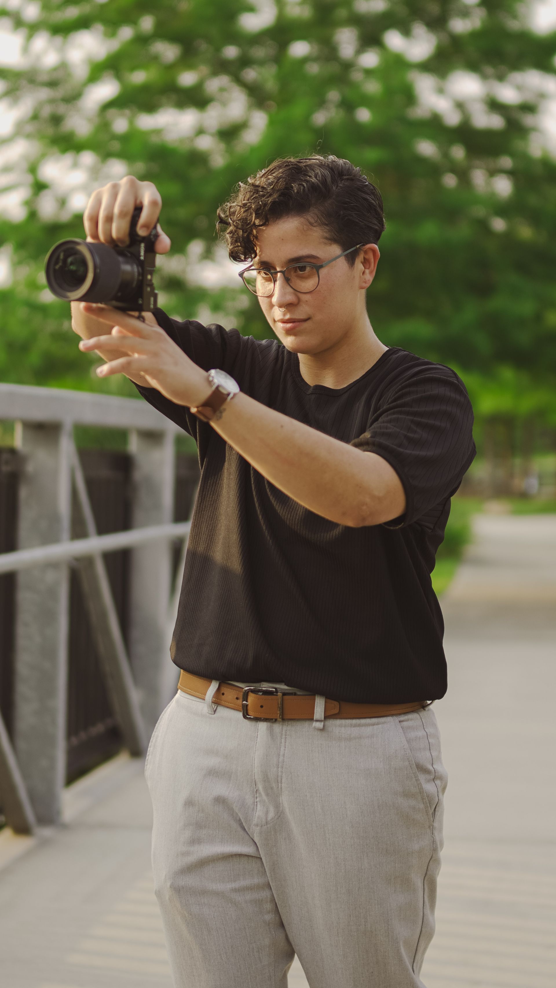Person holding a camera on a bridge, wearing glasses, black shirt, and light pants.