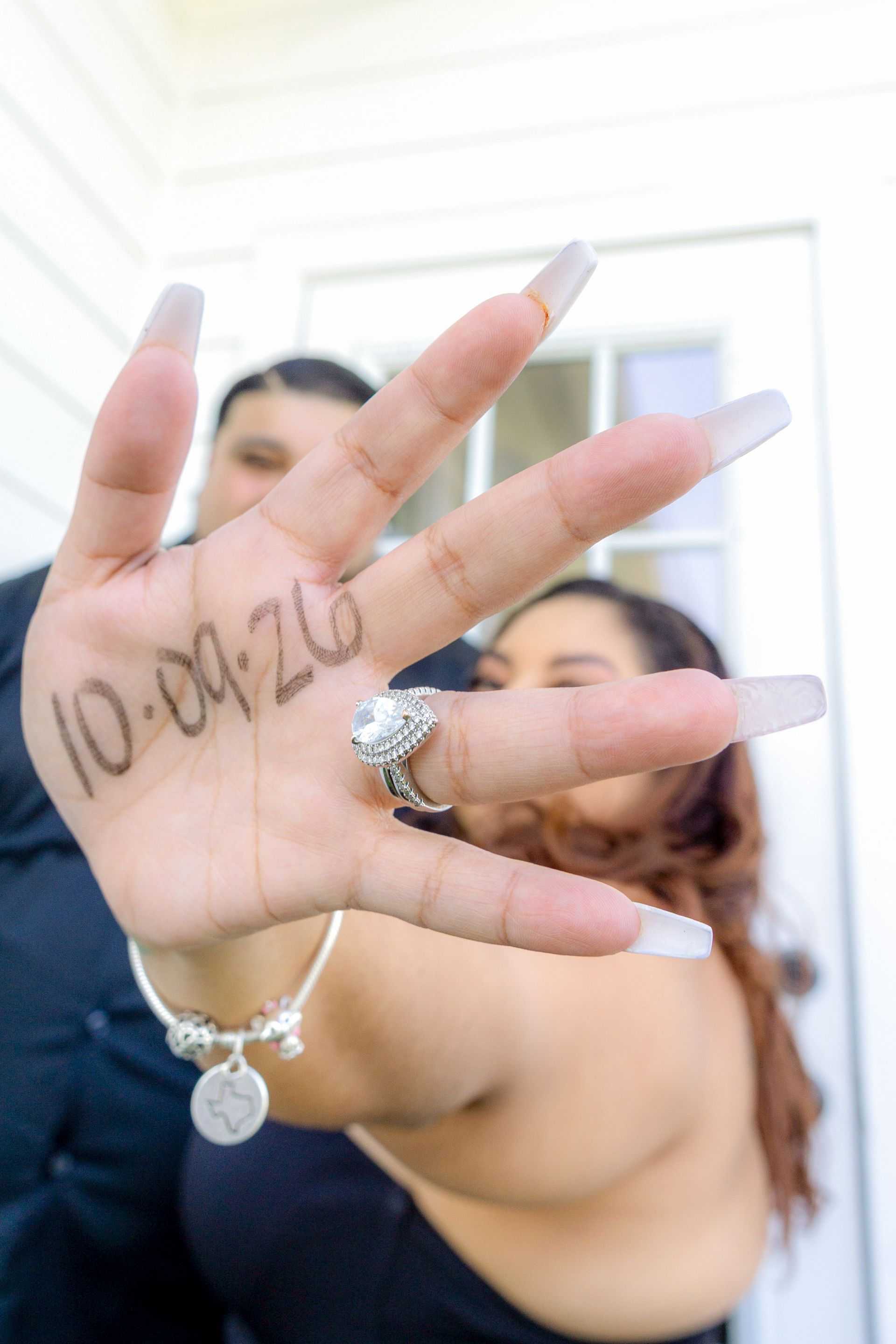 Hand with long white nails and ring showing a tattoo on the palm, held in front of a blurred mirror selfie.
