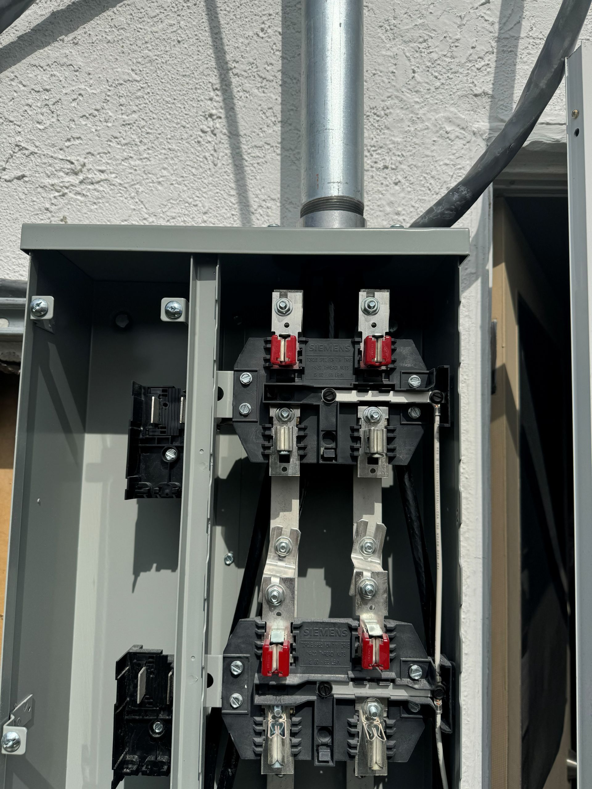 Gray electrical panel with exposed metal contacts, mounted on a building exterior. Conduit and cable visible.