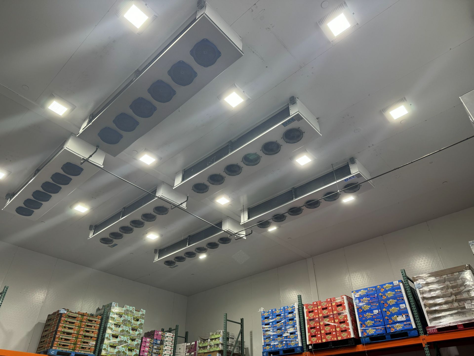 Warehouse ceiling with cooling units and square lights above stocked shelves.