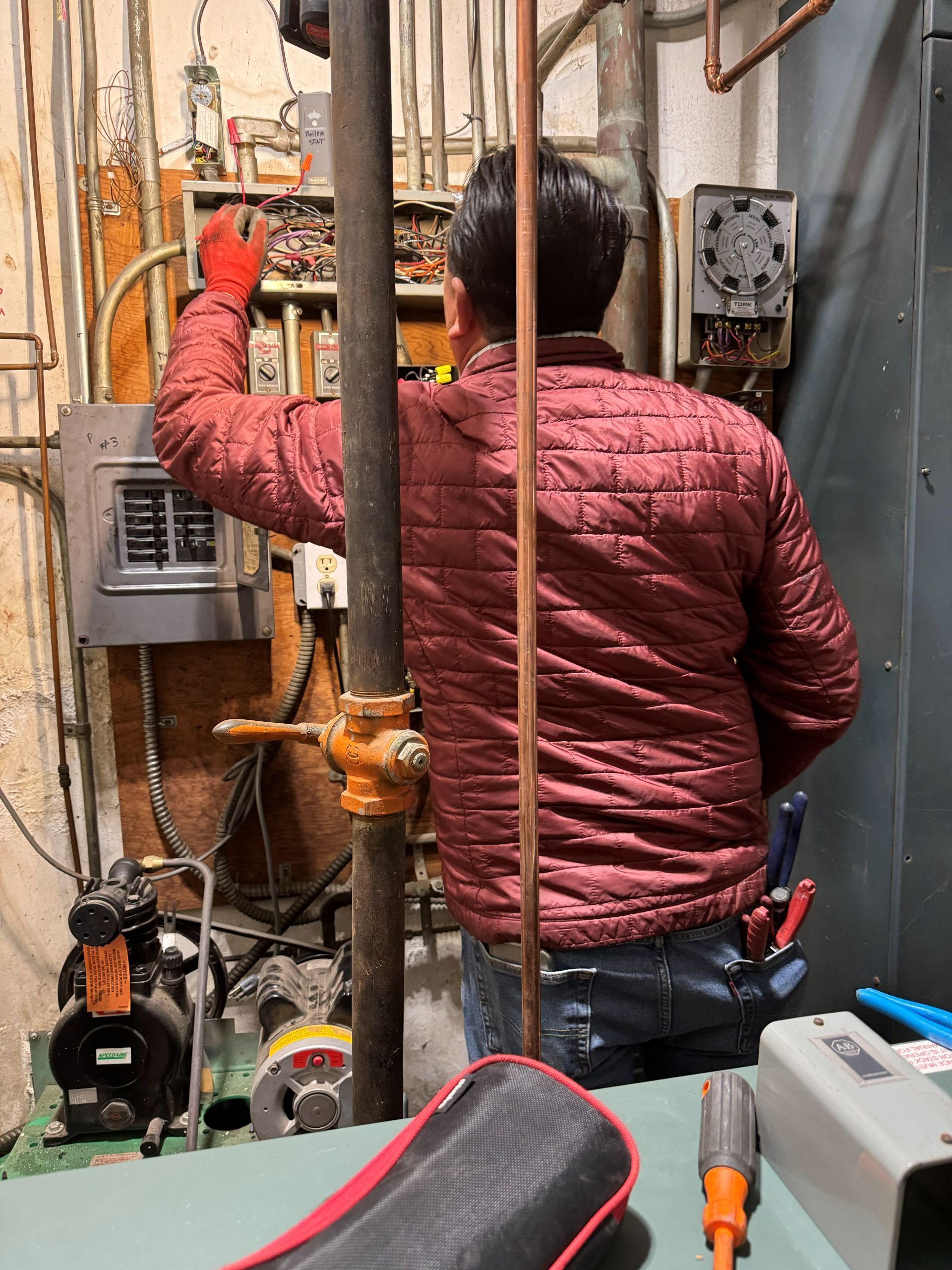 Person in red jacket working on electrical panel near pipes and machinery.