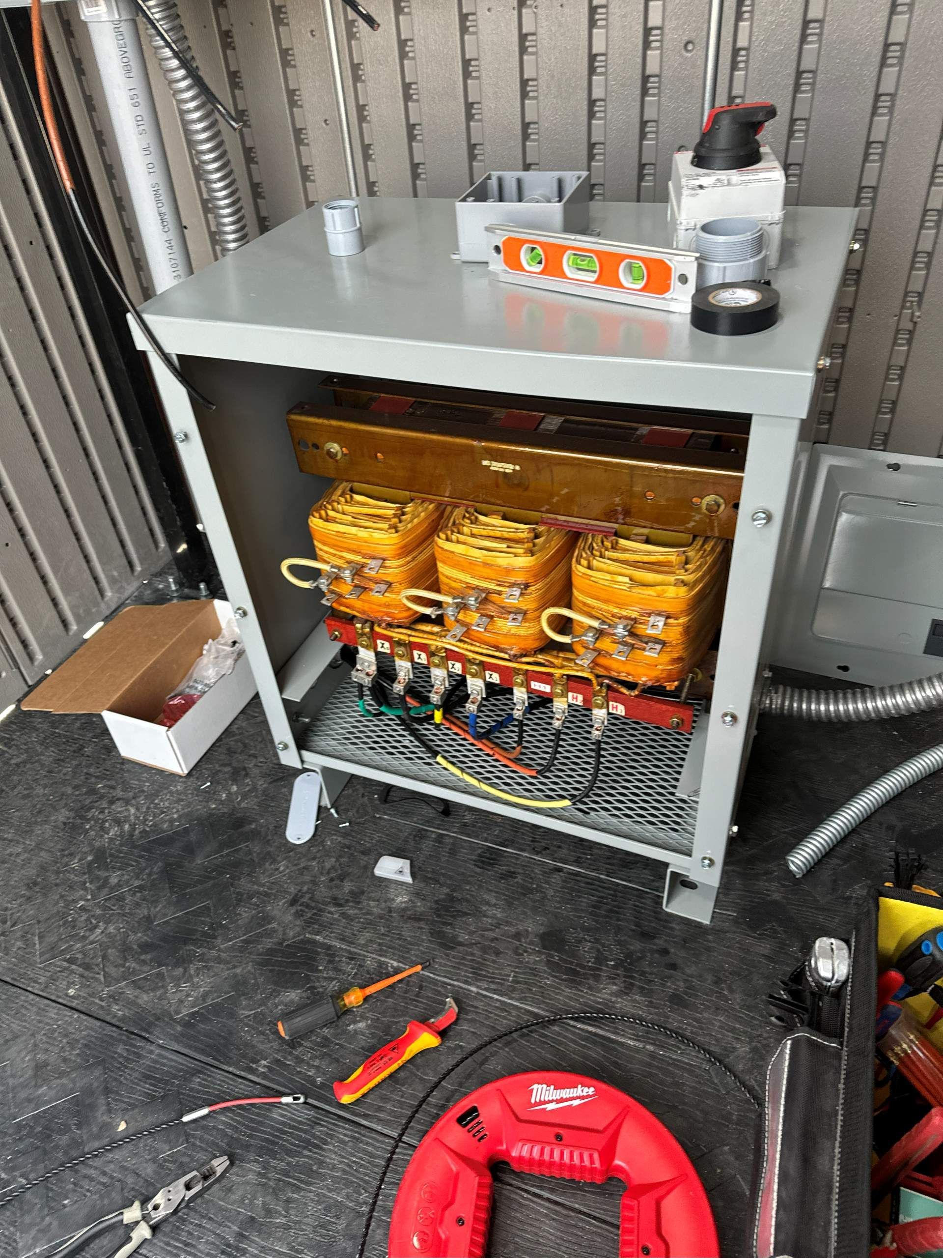 Open electrical transformer cabinet with wiring, tools, and construction materials on the floor.