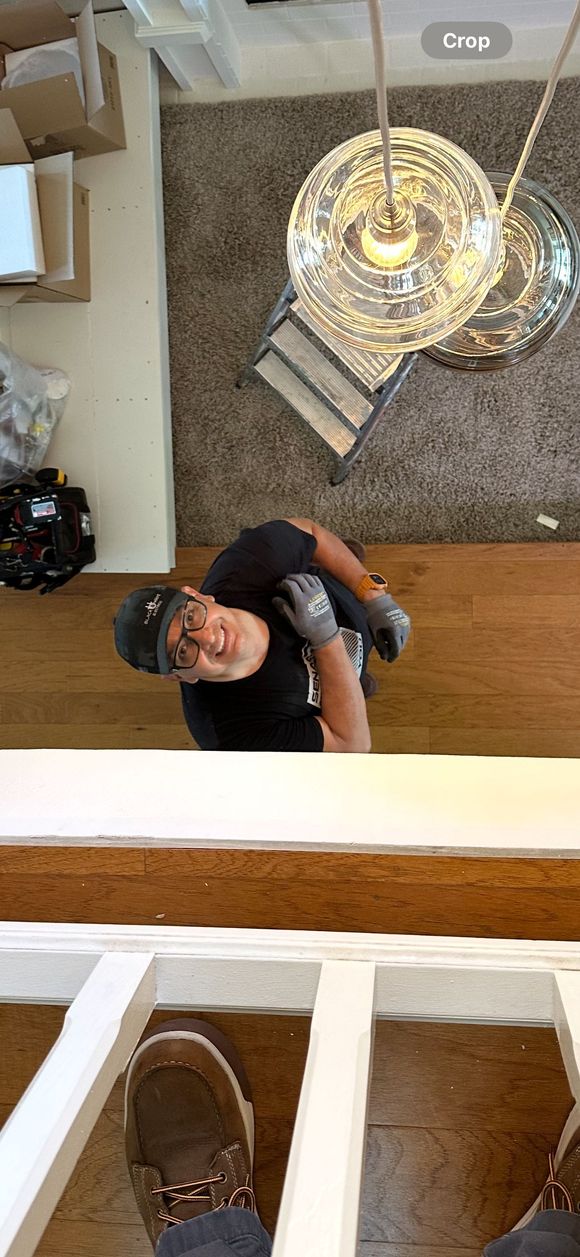 A man in a black shirt and cap looks up from below. He wears work gloves. A staircase and hanging lights are visible.
