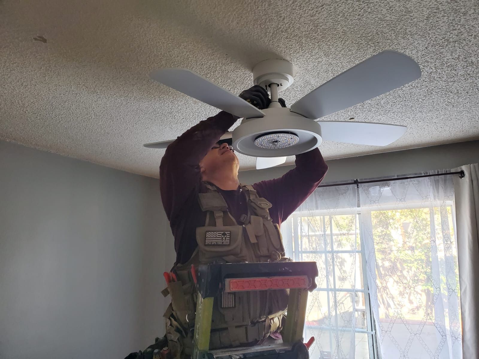 Person in a tactical vest installs a ceiling fan on a ladder in a room with white walls and a window.