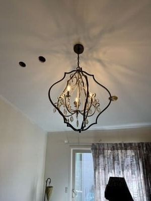 Ornate, dark metal chandelier hanging from a white ceiling, with a window and curtain visible below.