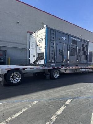 Generator in shipping container on flatbed truck, parked near a building on a sunny day.