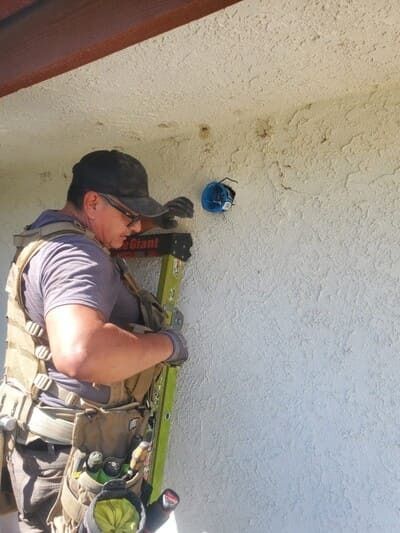 Person on a ladder, installing something into a blue electrical box on an exterior stucco wall.