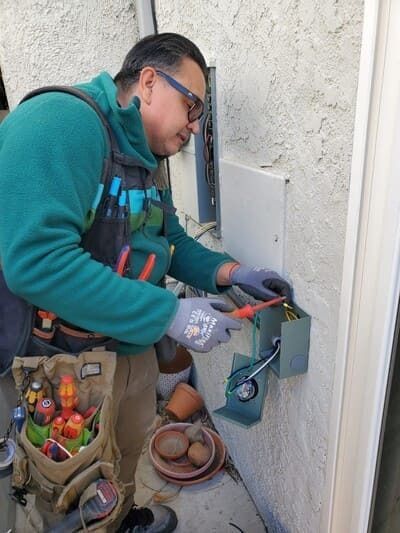 Electrician works on wiring in an outdoor electrical box; wearing gloves and a teal fleece.