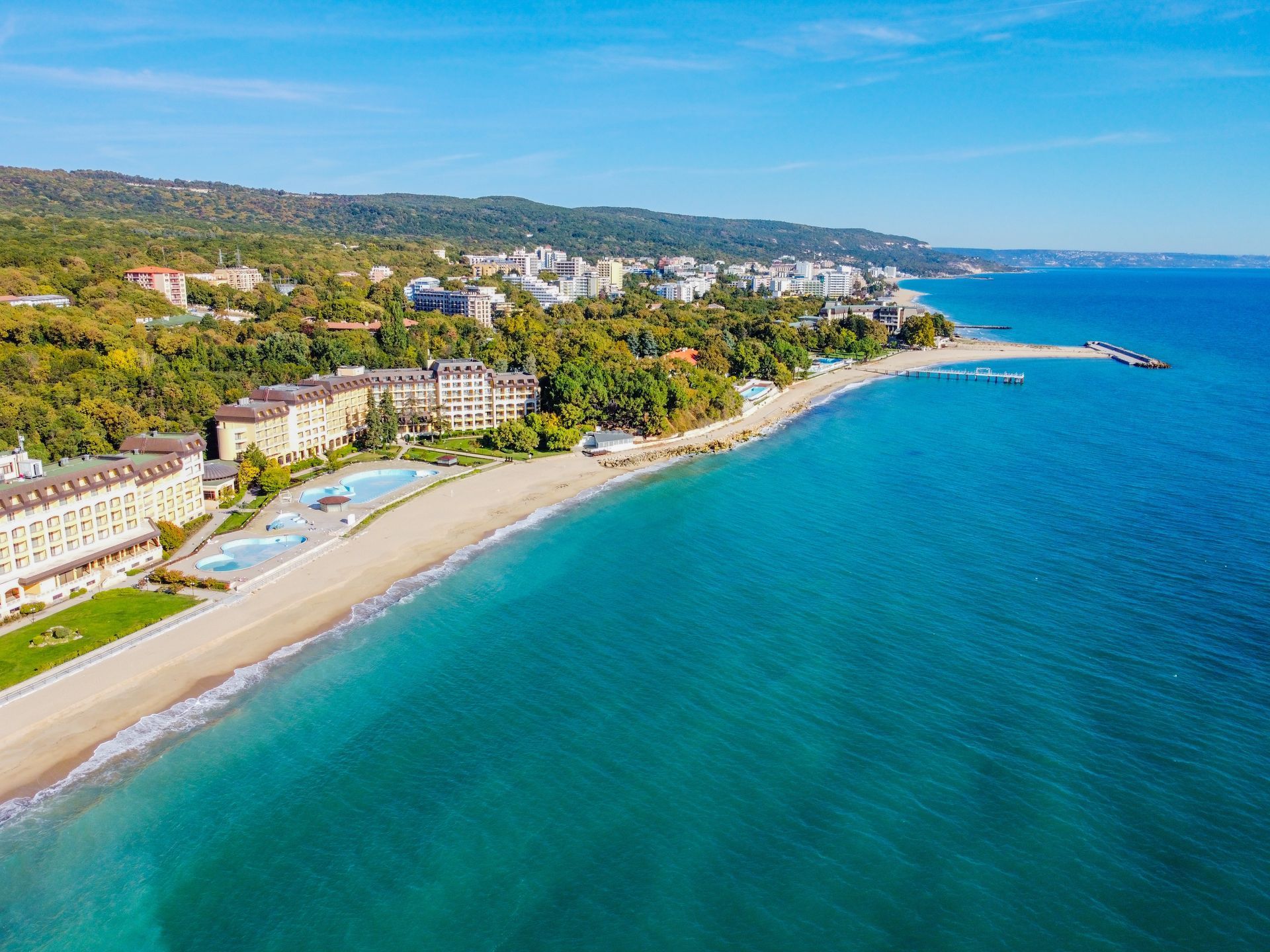 Aerial view of a coastal city with a turquoise sea, sandy beach, hotels, and a tree-covered hillside.