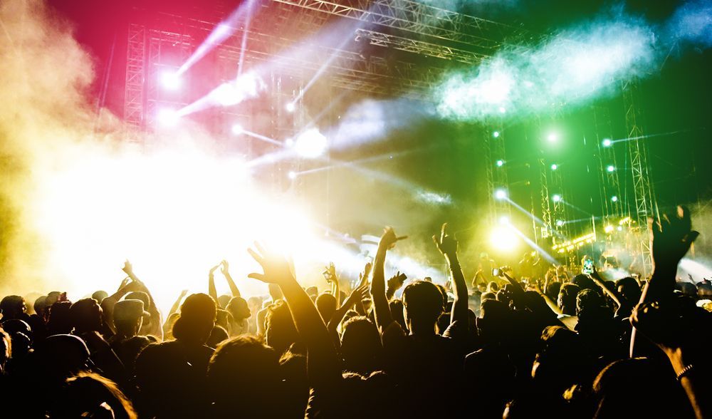 Concert crowd with arms raised toward a brightly lit stage with colorful lights and smoke.