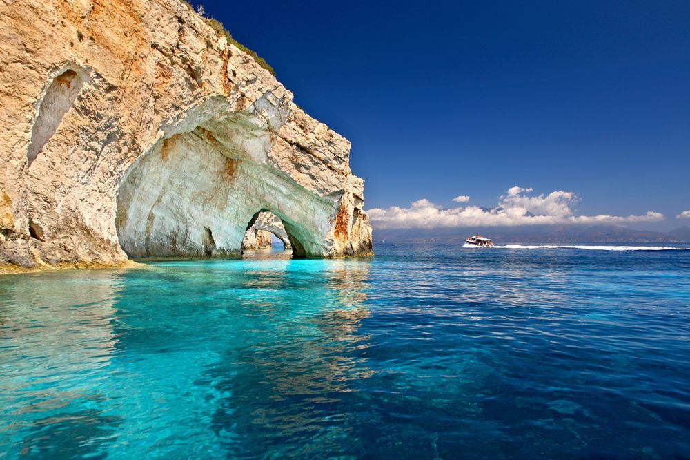 Cliffside caves over turquoise water with a boat on the horizon under a bright blue sky.