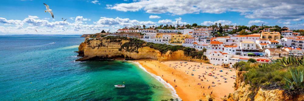 Coastal town with a beach and turquoise water under a blue sky. People sunbathe on the sand.