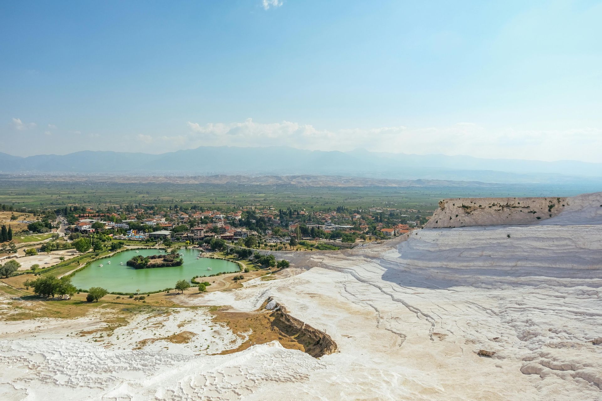 Pamukkale, Turkey: White travertine terraces overlooking a green lake and town against a backdrop of distant mountains.