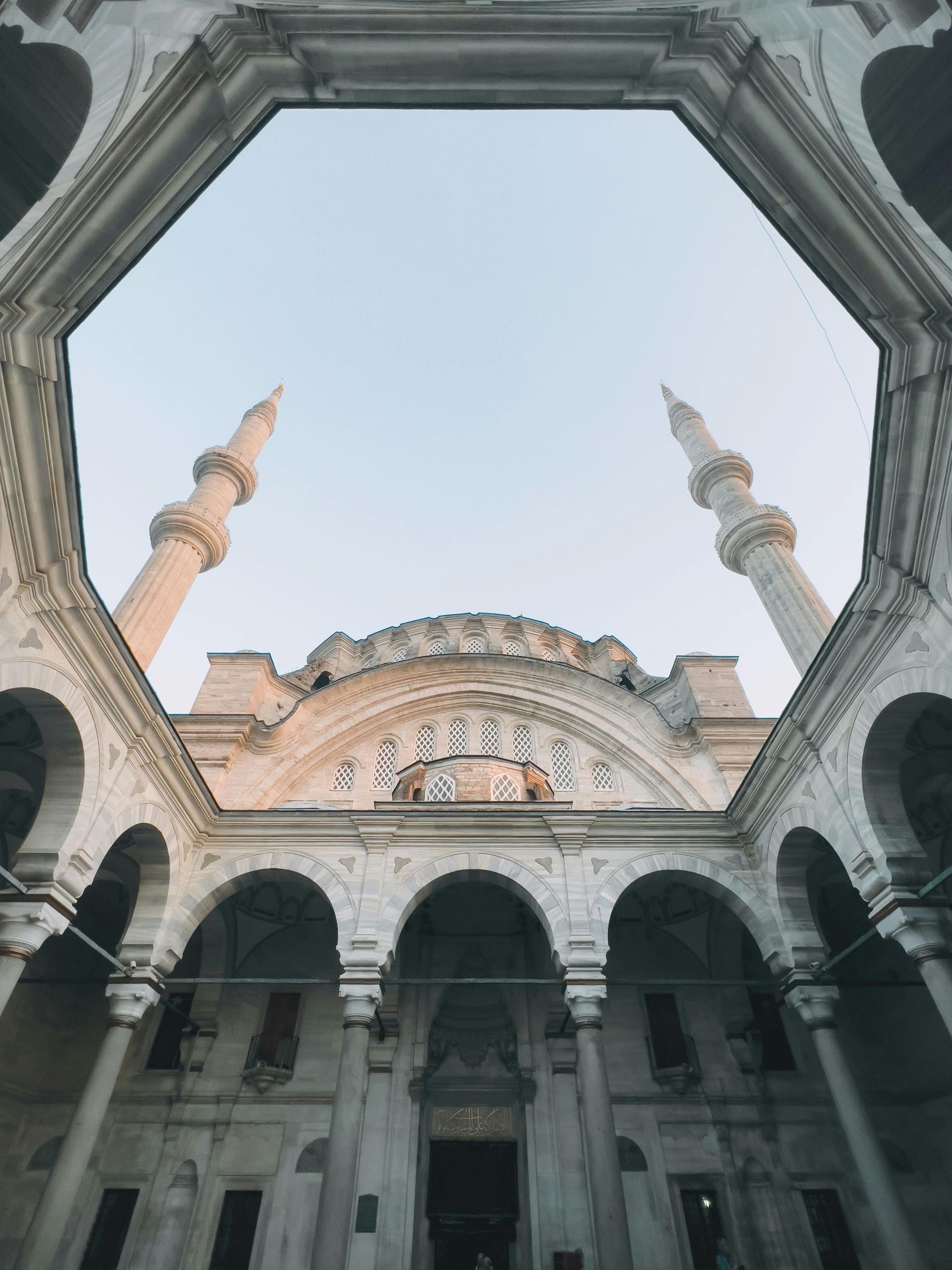 Low-angle view of a mosque courtyard with white archways, two minarets, and a pale blue sky.