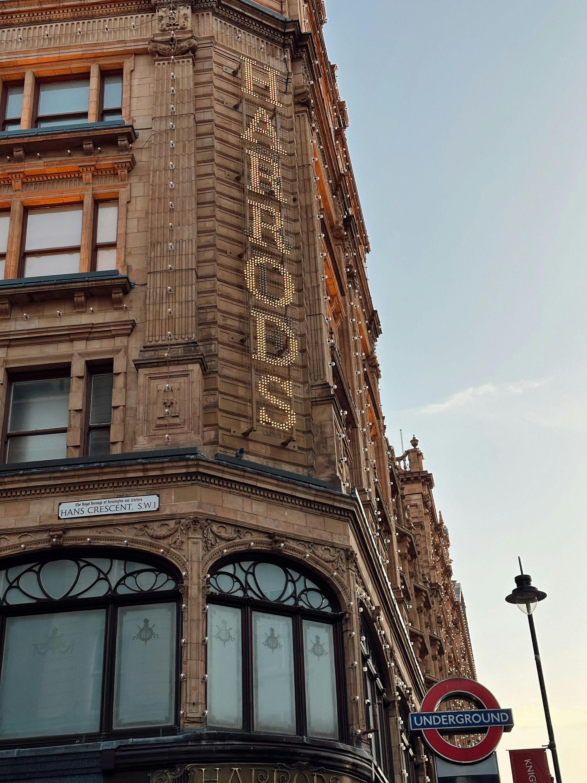Harrods department store exterior with illuminated sign against a pale blue sky; London Underground sign visible.